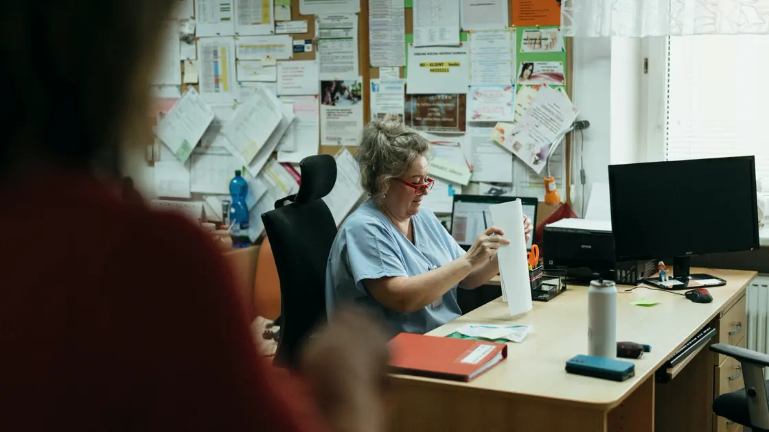 A nurse in blue scrubs at an office desk on a hospital ward, sorting paperwork