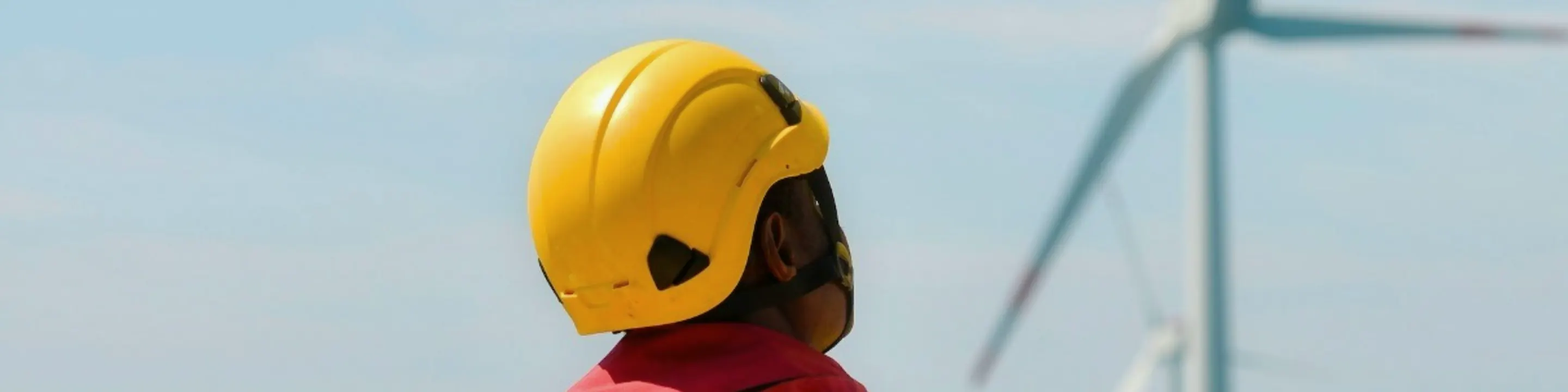 A service technician in a yellow hard hat, a wind turbine behind him